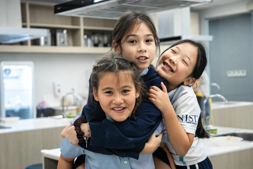 Three schoolchildren smiling and hugging in a classroom, showing friendship and a sense of belonging at school. Image via NIST