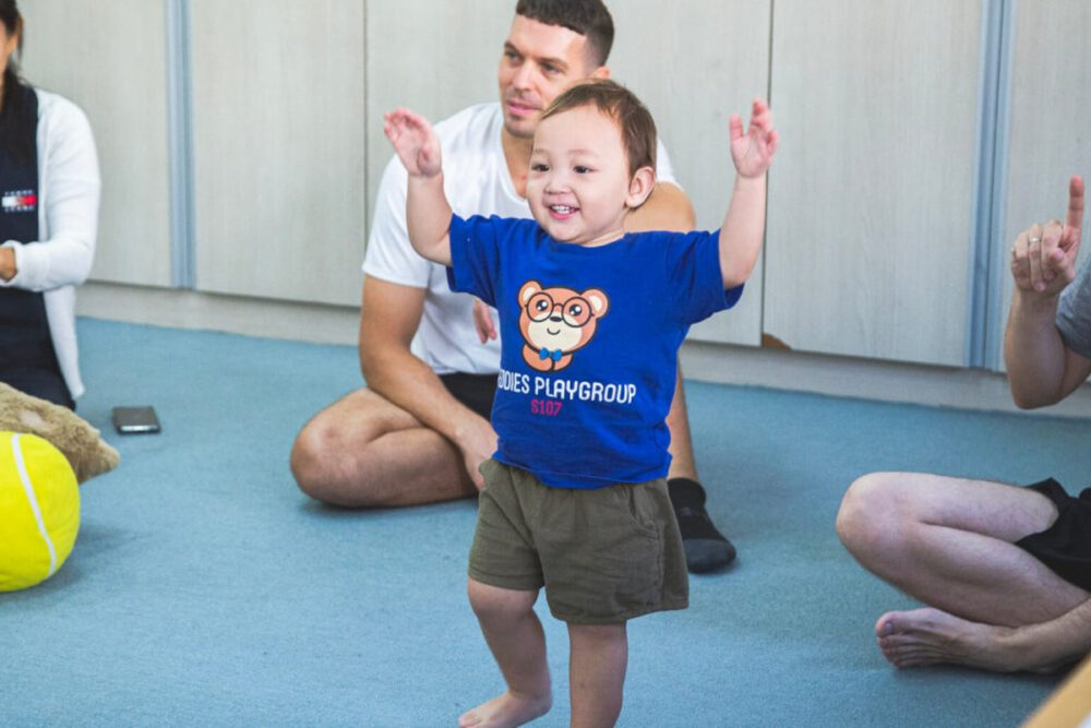 A toddler smiles and raises their arms during a music and movement session, surrounded by parents and young children in a playgroup. Image via St. Andrews International School