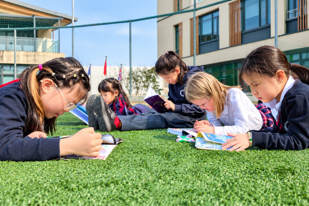 Schoolgirls reading books and studying together on the grass at SPGS International School, enjoying an outdoor learning activity. Image via The BSA
